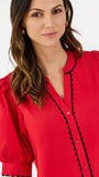 A woman modeling a bright red short-sleeve blouse with black scalloped trim along the button placket and sleeve edges, paired with white pants, against a plain white studio background.