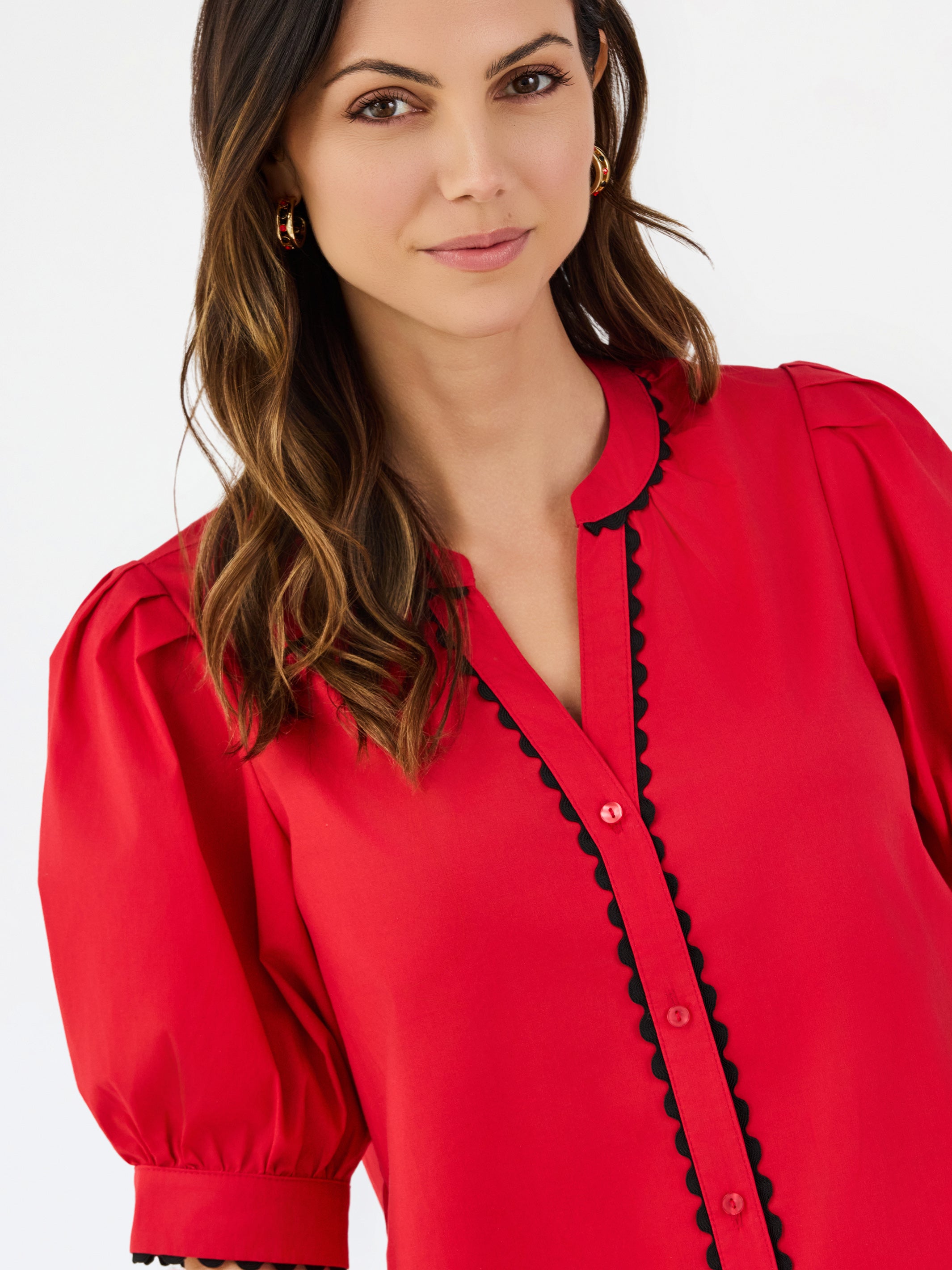 A woman modeling a bright red short-sleeve blouse with black scalloped trim along the button placket and sleeve edges, paired with white pants, against a plain white studio background.