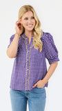 A woman modeling a purple and white gingham short-sleeve button-up blouse with gold trim, paired with blue jeans, against a plain white studio background.