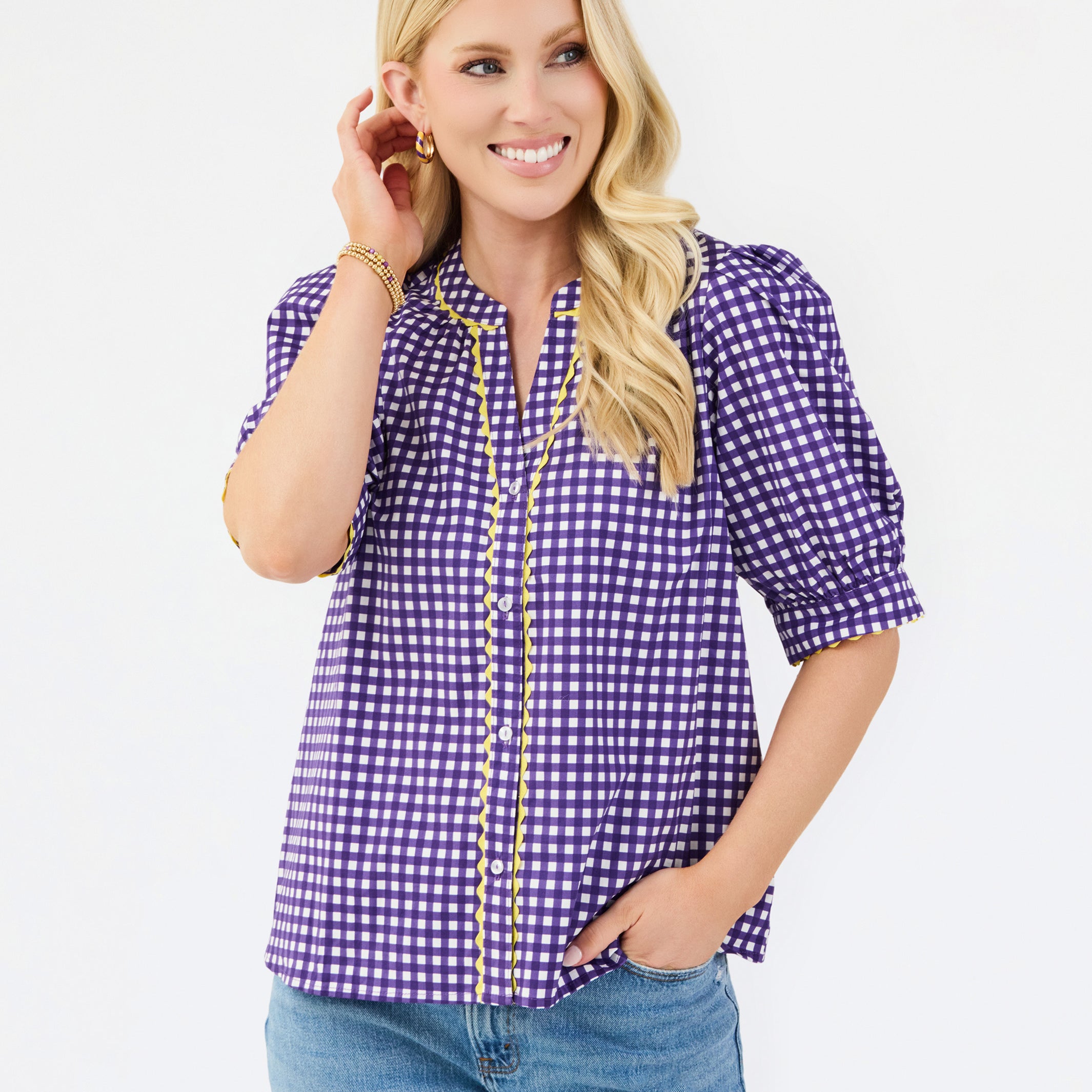 A woman modeling a purple and white gingham short-sleeve button-up blouse with gold trim, paired with blue jeans, against a plain white studio background.