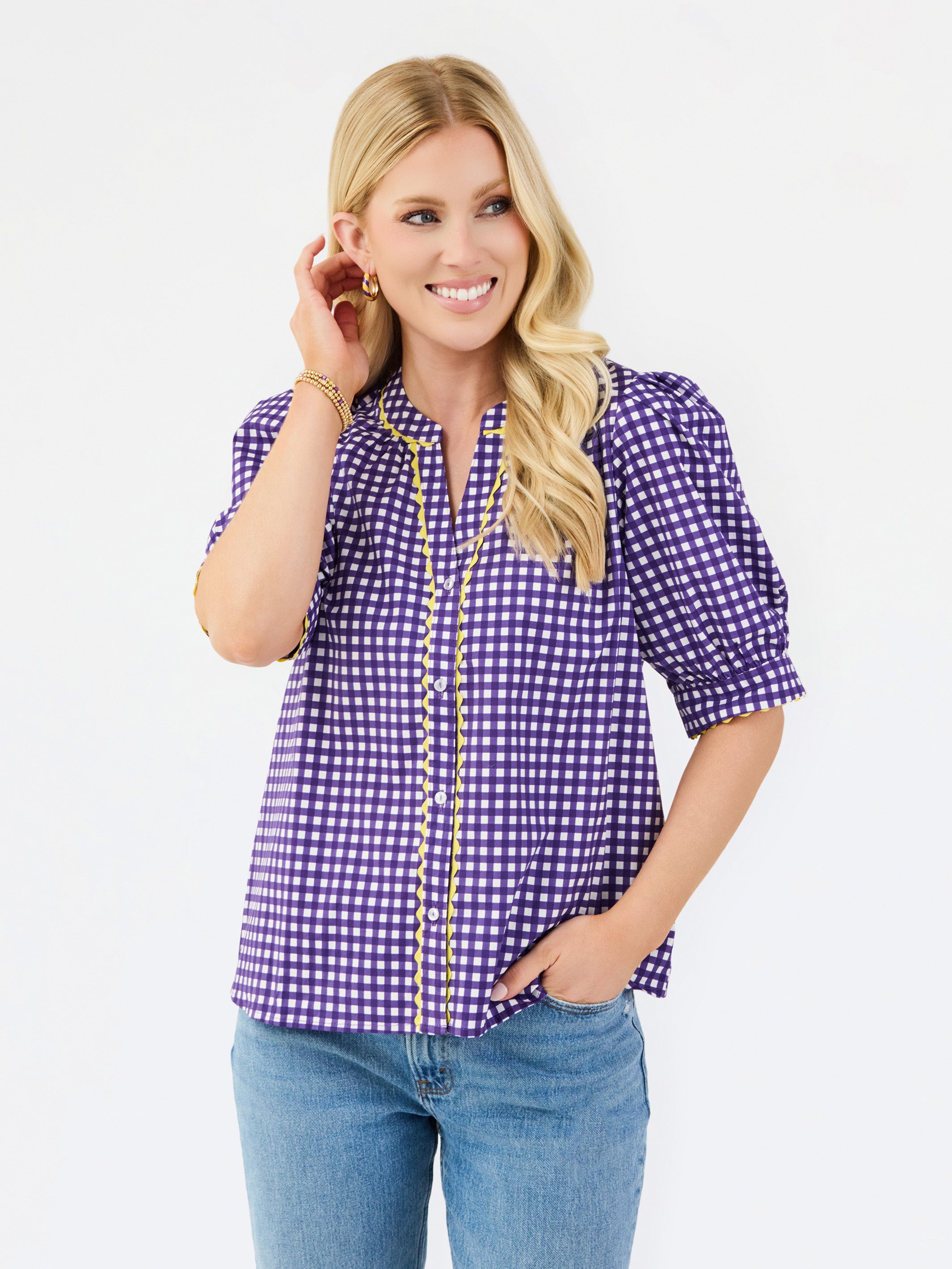 A woman modeling a purple and white gingham short-sleeve button-up blouse with gold trim, paired with blue jeans, against a plain white studio background.