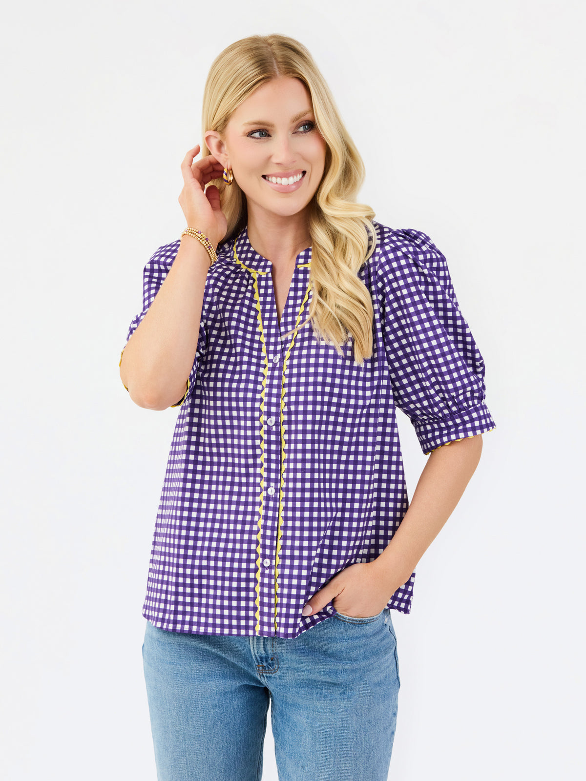 A woman modeling a purple and white gingham short-sleeve button-up blouse with gold trim, paired with blue jeans, against a plain white studio background.