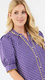 A blonde woman wearing a purple and white gingham short-sleeve blouse with yellow trim, photographed against a plain light gray studio background.