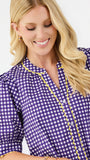A smiling blonde woman wearing a purple and white gingham blouse with yellow trim, photographed against a clean white studio background.