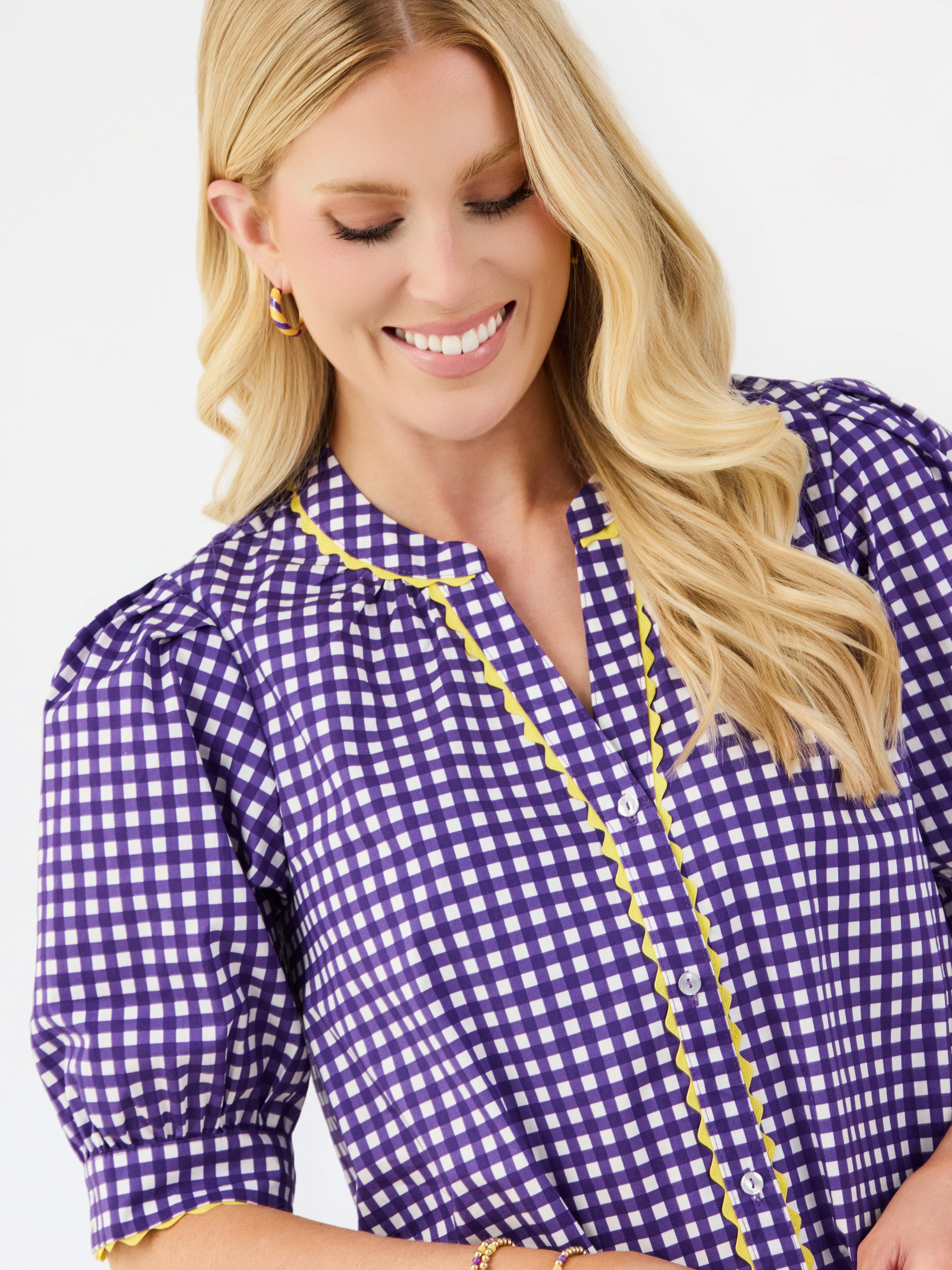 A smiling blonde woman wearing a purple and white gingham blouse with yellow trim, photographed against a clean white studio background.