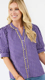 A smiling blonde woman wearing a purple and white gingham short-sleeve blouse with yellow ruffle trim along the split neckline and sleeve cuffs, paired with blue jeans against a light gray studio background.