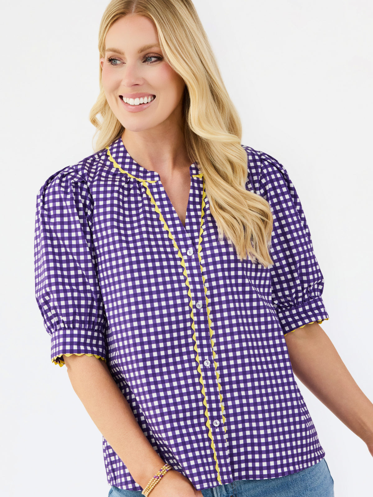 A smiling blonde woman wearing a purple and white gingham short-sleeve blouse with yellow ruffle trim along the split neckline and sleeve cuffs, paired with blue jeans against a light gray studio background.