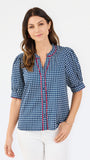 A woman modeling a short-sleeve button-up blouse with a blue and white gingham pattern and red piping details on the placket and sleeve cuffs, paired with white pants against a plain white studio background.