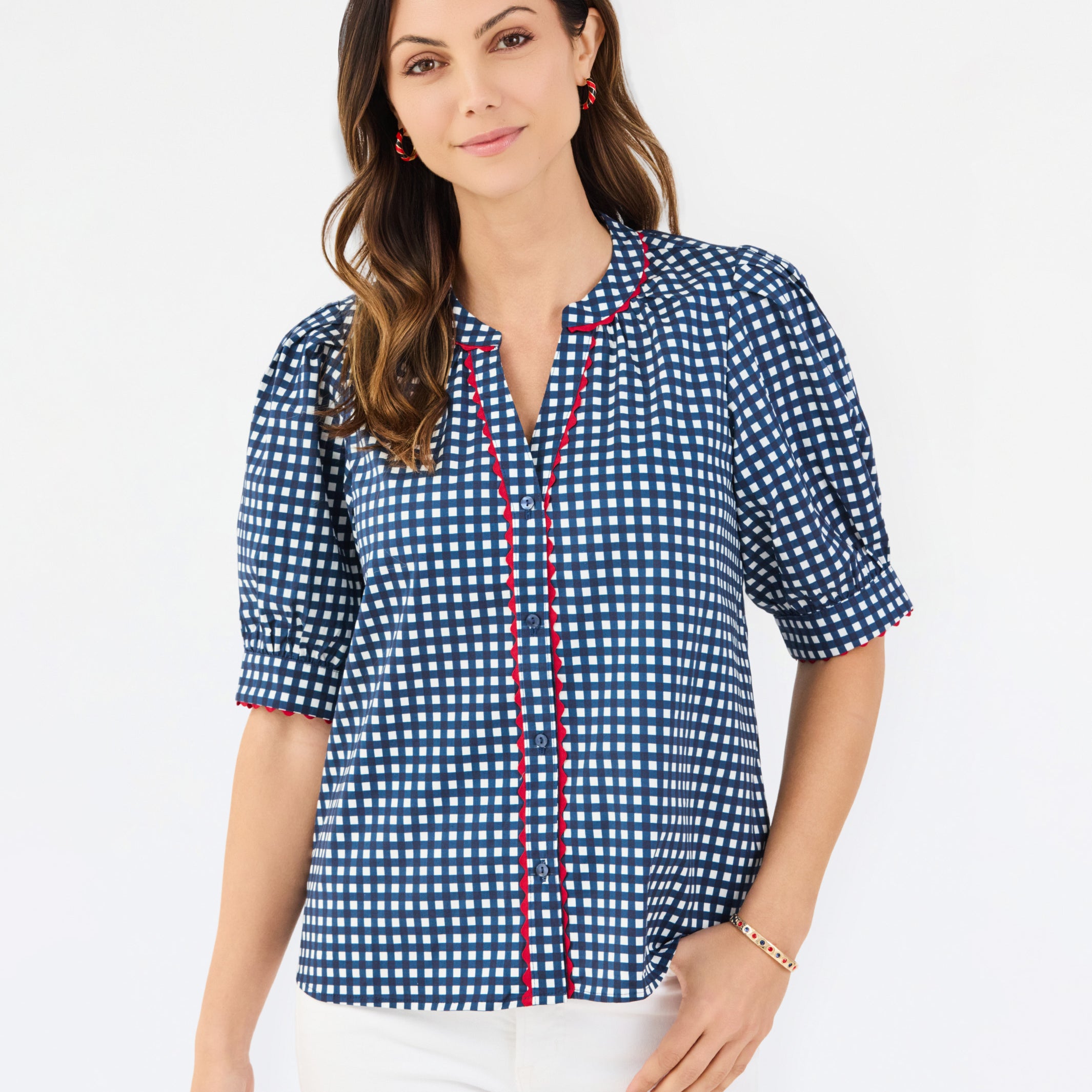 A woman modeling a short-sleeve button-up blouse with a blue and white gingham pattern and red piping details on the placket and sleeve cuffs, paired with white pants against a plain white studio background.