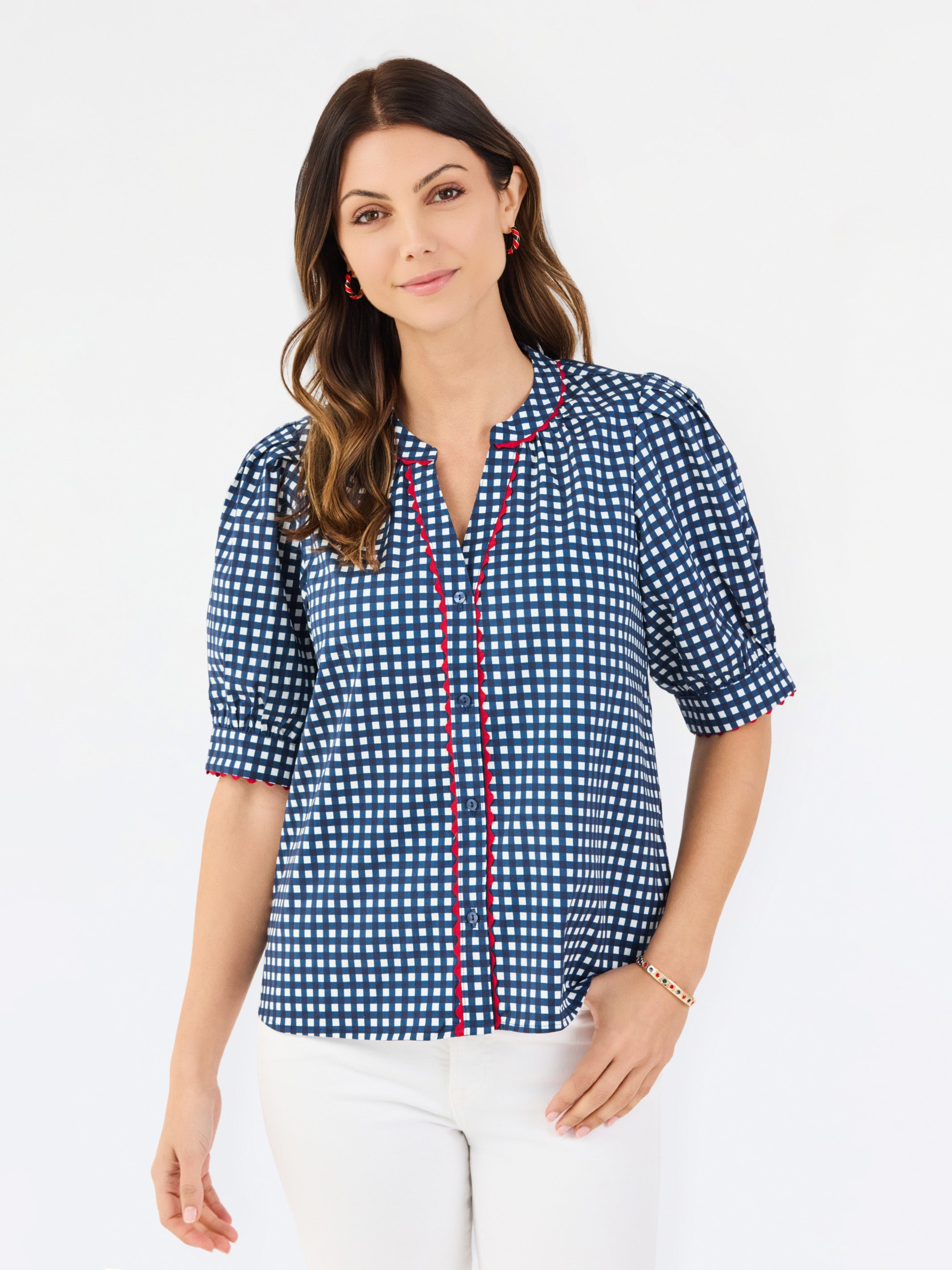 A woman modeling a short-sleeve button-up blouse with a blue and white gingham pattern and red piping details on the placket and sleeve cuffs, paired with white pants against a plain white studio background.