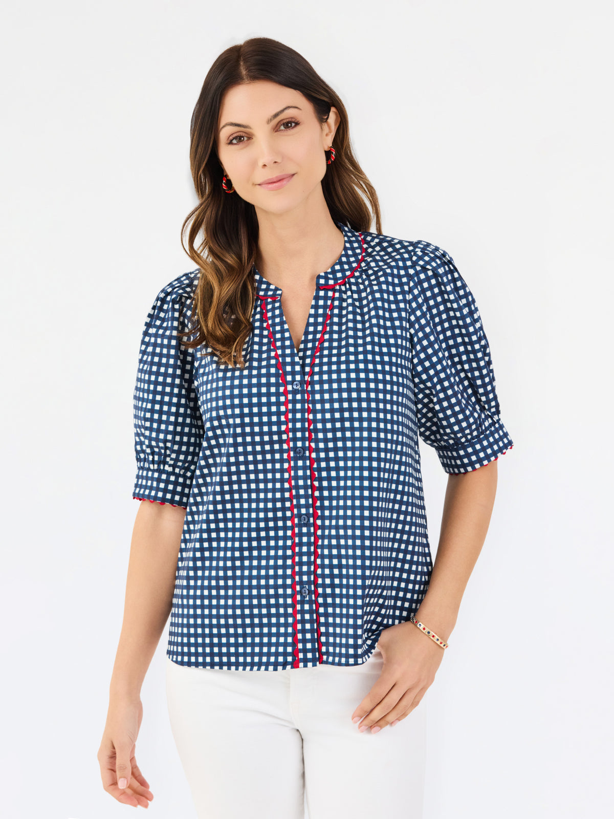A woman modeling a short-sleeve button-up blouse with a blue and white gingham pattern and red piping details on the placket and sleeve cuffs, paired with white pants against a plain white studio background.