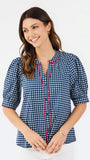 A woman modeling a short-sleeve button-up blouse with a blue and white gingham pattern and red piping details on the placket and sleeve cuffs, paired with white pants against a plain white studio background.