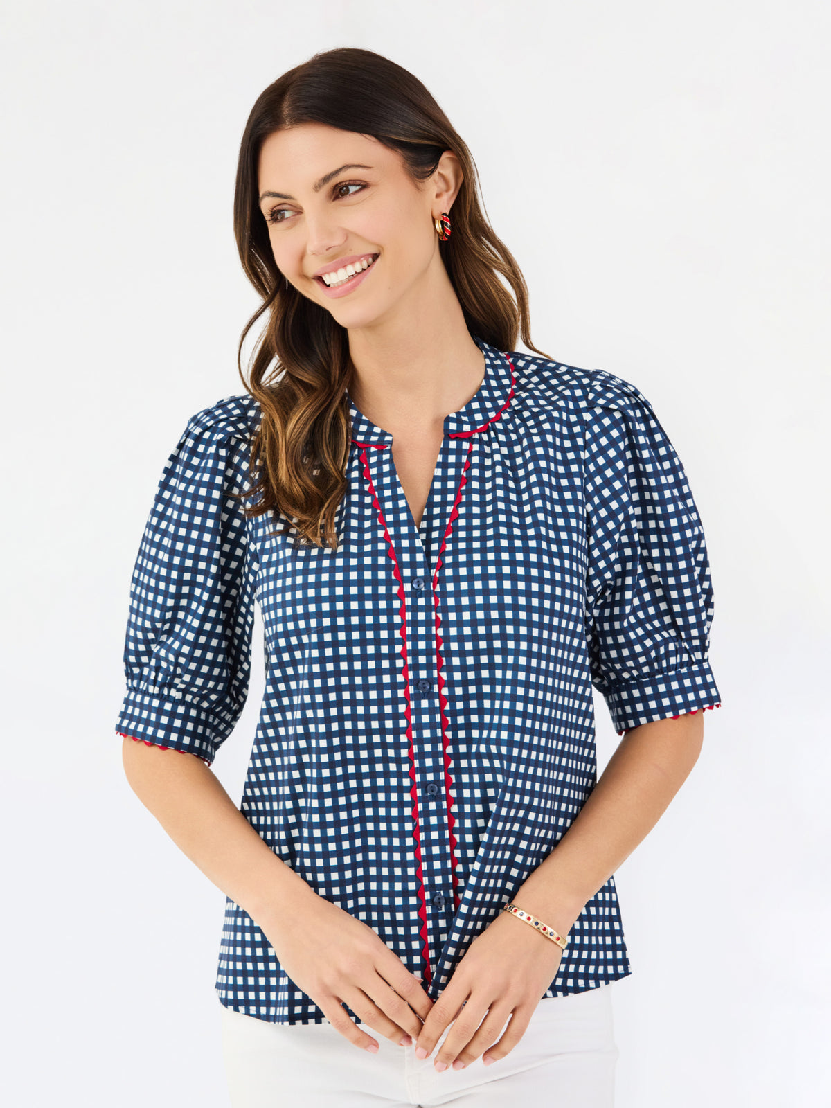 A woman modeling a short-sleeve button-up blouse with a blue and white gingham pattern and red piping details on the placket and sleeve cuffs, paired with white pants against a plain white studio background.
