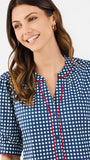 A woman modeling a short-sleeve button-up blouse with a blue and white gingham pattern and red piping details on the placket and sleeve cuffs, paired with white pants against a plain white studio background.