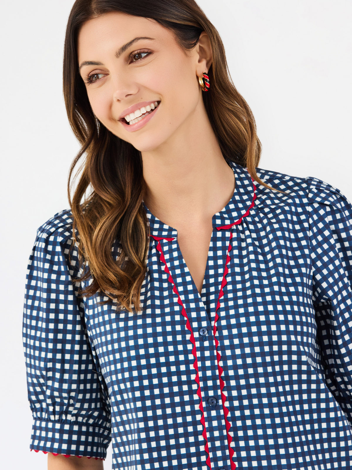 A woman modeling a short-sleeve button-up blouse with a blue and white gingham pattern and red piping details on the placket and sleeve cuffs, paired with white pants against a plain white studio background.