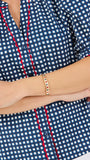 A woman modeling a short-sleeve button-up blouse with a blue and white gingham pattern and red piping details on the placket and sleeve cuffs, paired with white pants against a plain white studio background.
