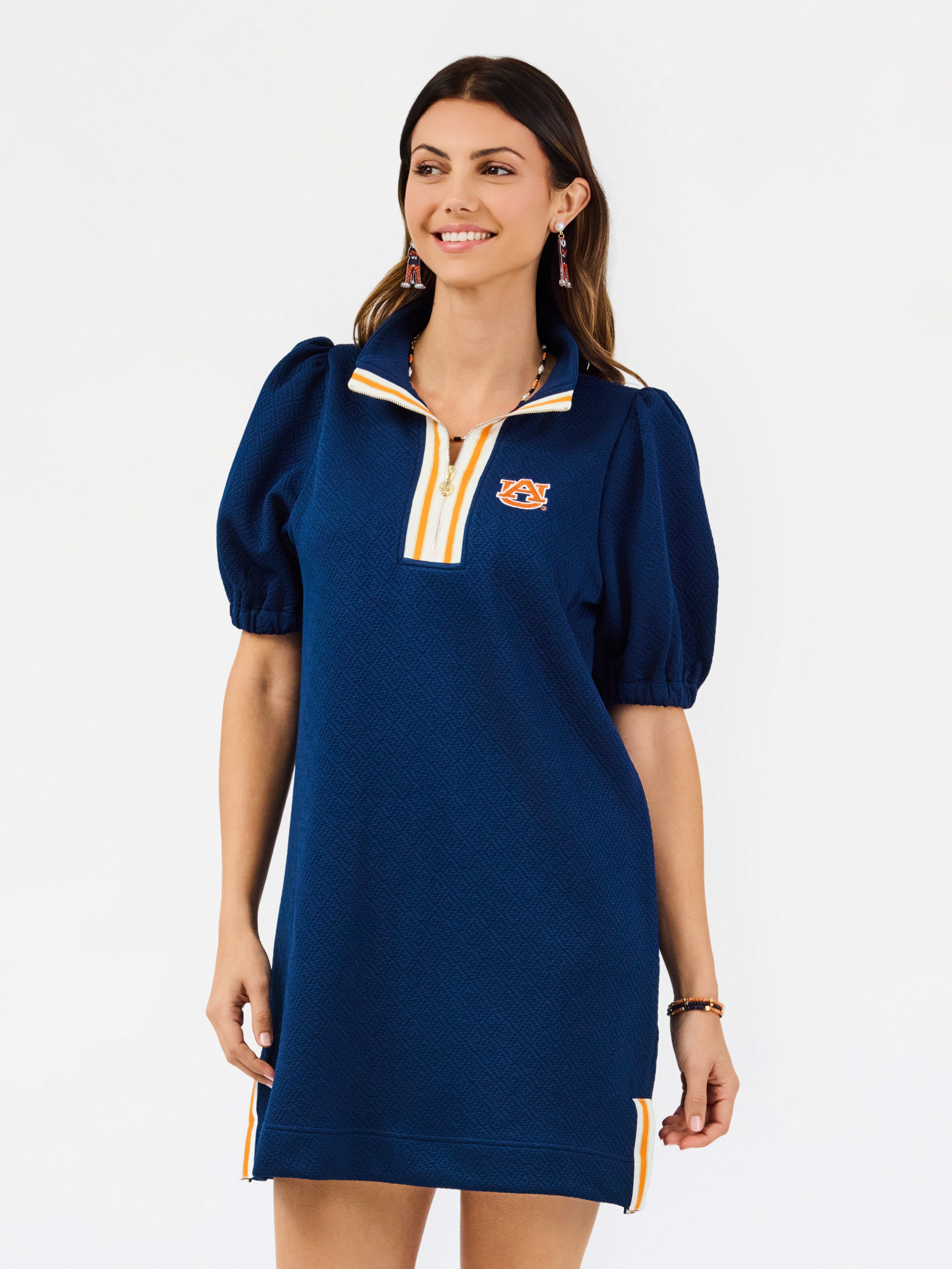 A woman modeling a navy blue short-sleeve polo dress with gold and white trim on the collar and hem, featuring a small embroidered tiger logo on the left chest.