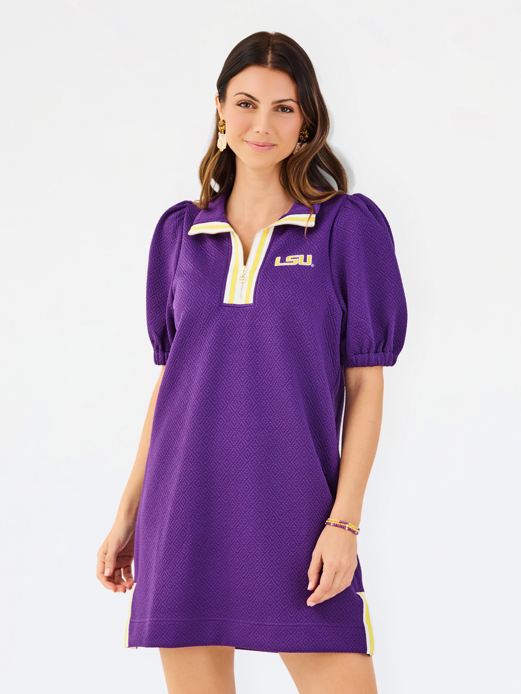 A woman modeling a purple LSU athletic polo dress with puff sleeves, a half-zip collar, and a gold and white LSU logo on the chest, set against a plain white studio background.