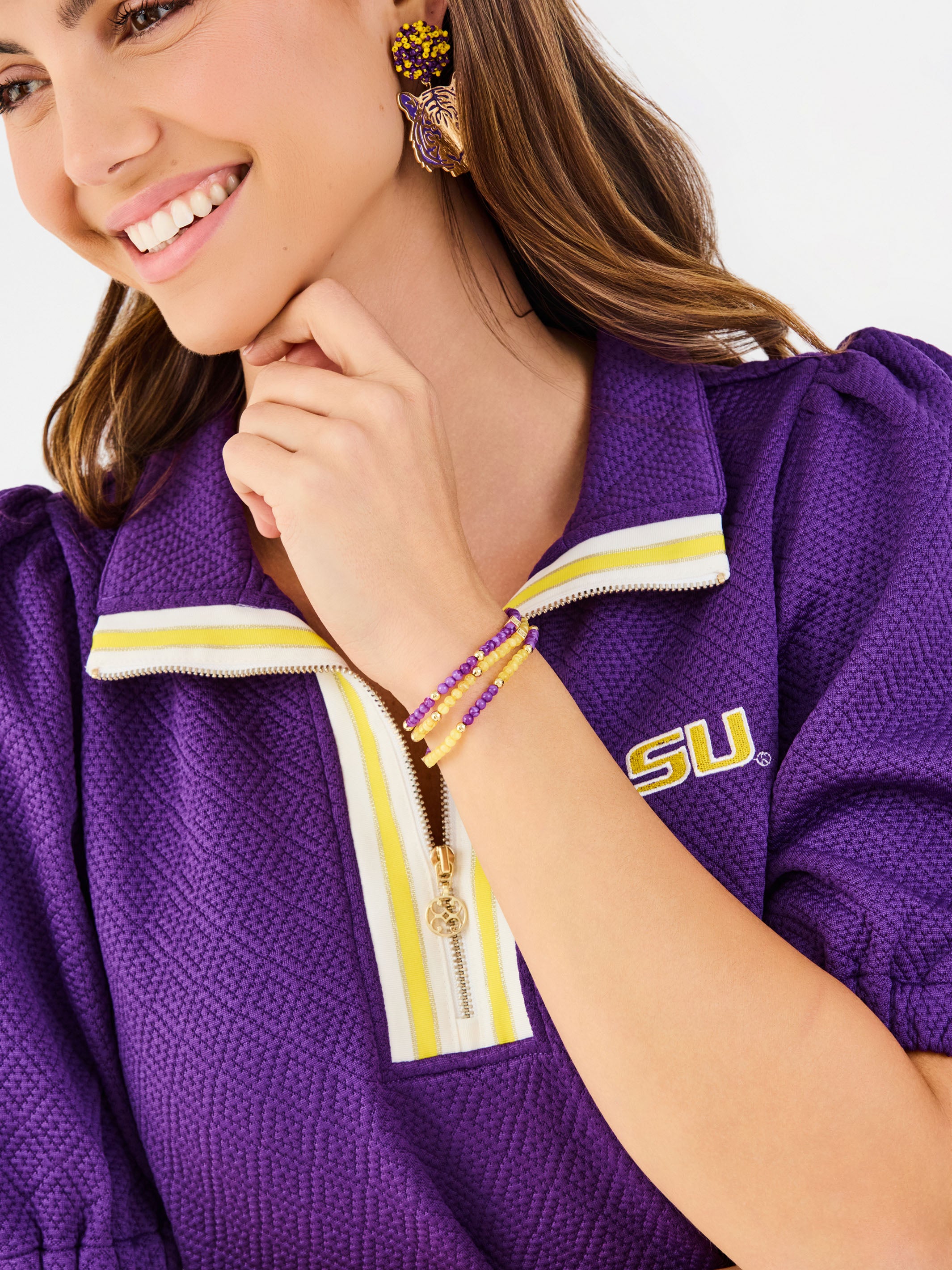 A close-up lifestyle shot of a woman wearing a purple LSU athletic quarter-zip pullover. The pullover features a textured purple mesh fabric, a white and yellow striped collar, a gold zipper, and a yellow 'LSU' logo on the chest.
