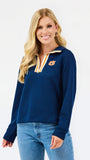 A smiling blonde woman wearing a navy blue quarter-zip sweatshirt with a gold and white striped collar trim and a gold tiger logo on the left chest, paired with blue jeans, against a plain white studio background.