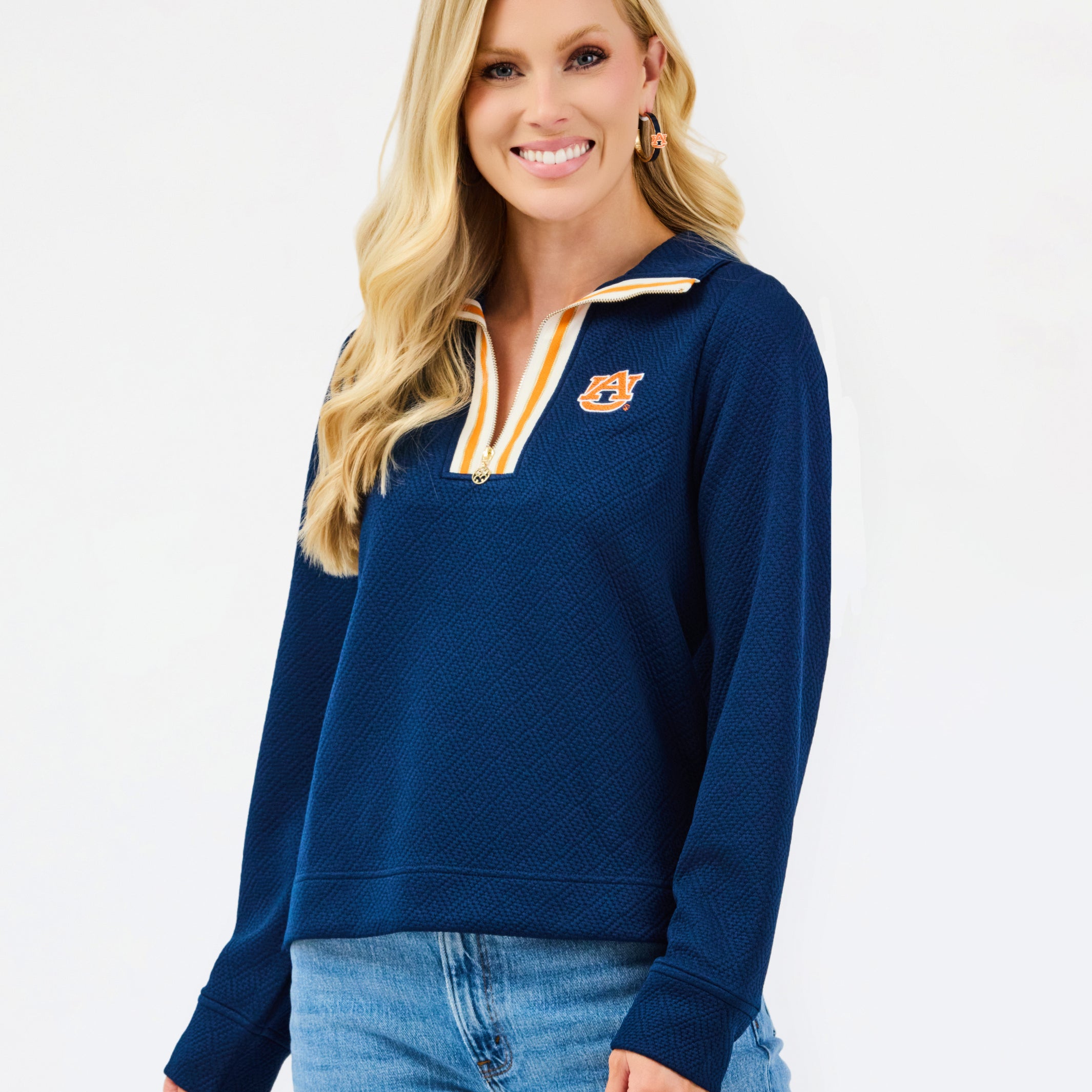A smiling blonde woman wearing a navy blue quarter-zip sweatshirt with a gold and white striped collar trim and a gold tiger logo on the left chest, paired with blue jeans, against a plain white studio background.