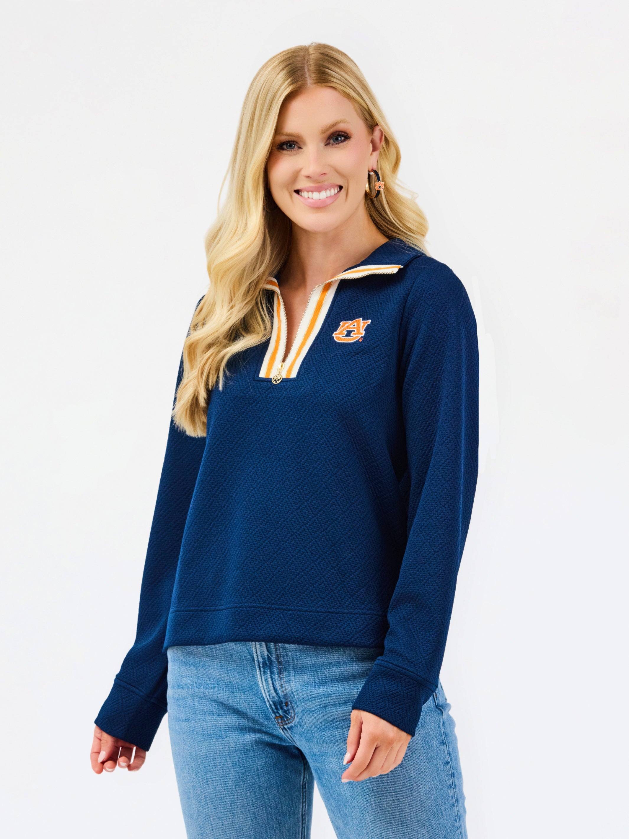 A smiling blonde woman wearing a navy blue quarter-zip sweatshirt with a gold and white striped collar trim and a gold tiger logo on the left chest, paired with blue jeans, against a plain white studio background.