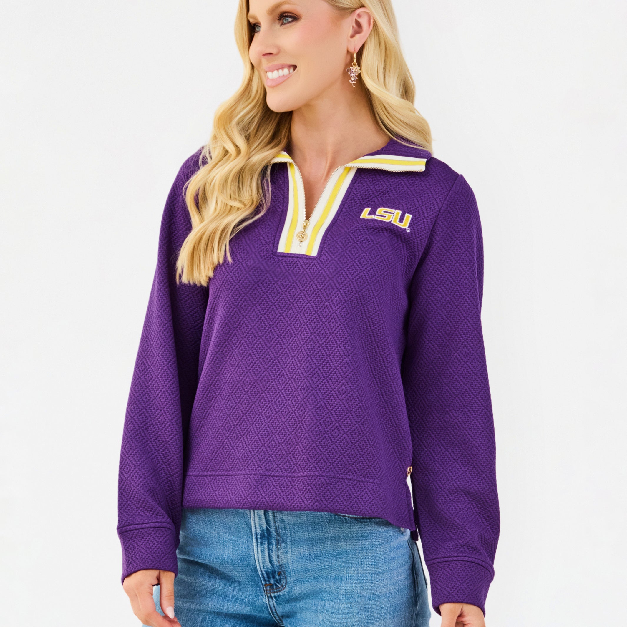 A woman wearing a purple LSU quarter-zip pullover sweatshirt with yellow and white trim, paired with blue jeans, against a plain white studio background.
