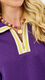 Close-up of a woman wearing a purple knit top with a white and yellow striped collar, layered with a purple and yellow beaded necklace and a gold coin pendant.