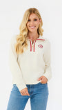 A woman modeling a cream-colored quarter-zip sweatshirt with red trim and a circular red and white logo on the left chest, paired with blue jeans against a plain white background.