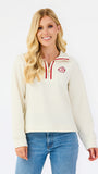 A woman modeling a cream-colored quarter-zip sweatshirt with red trim and a circular red and white logo on the left chest, paired with blue jeans against a plain white background.