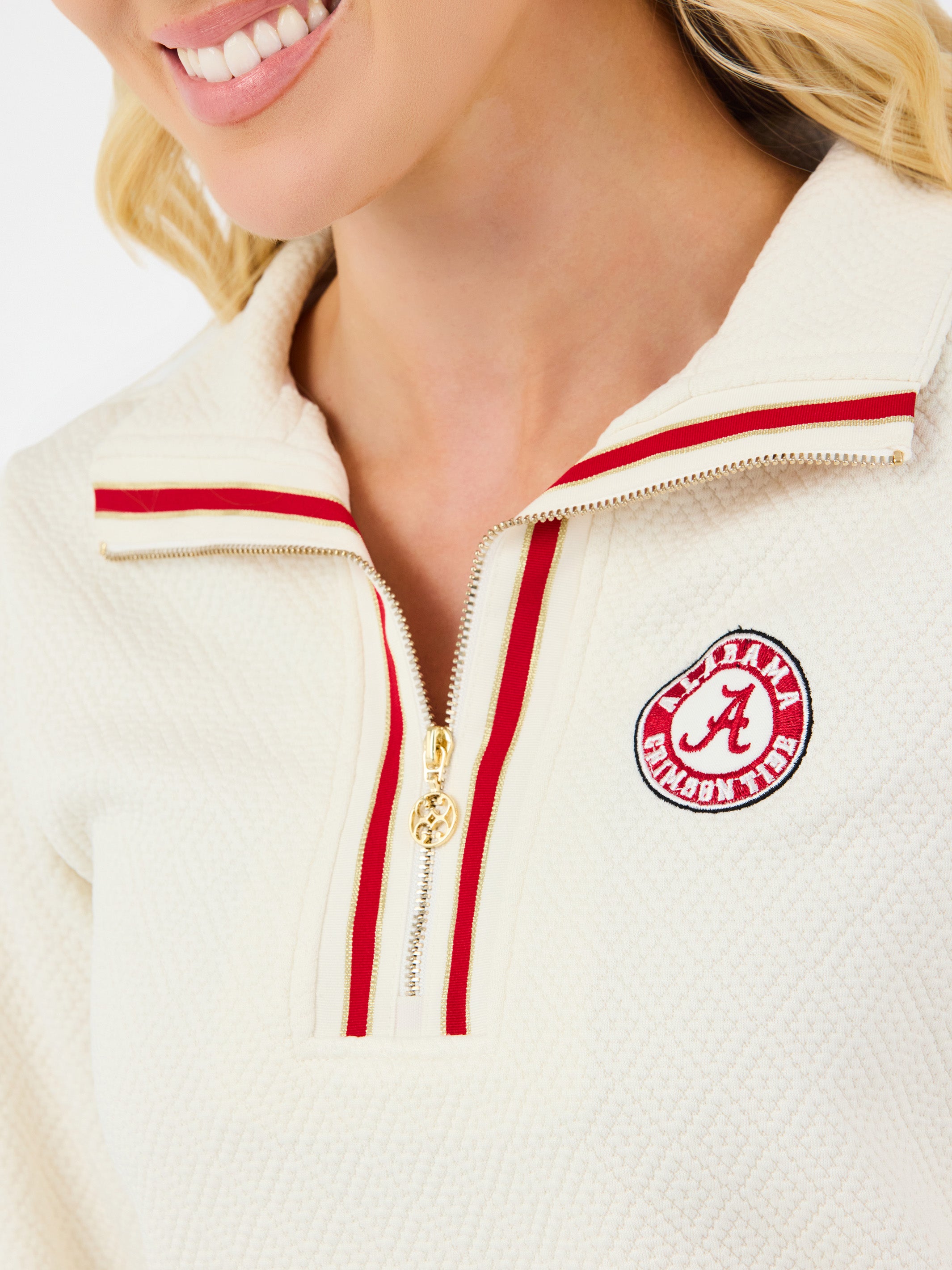 A woman modeling a cream-colored quarter-zip sweatshirt with red trim and a circular red and white logo on the left chest, paired with blue jeans against a plain white background.