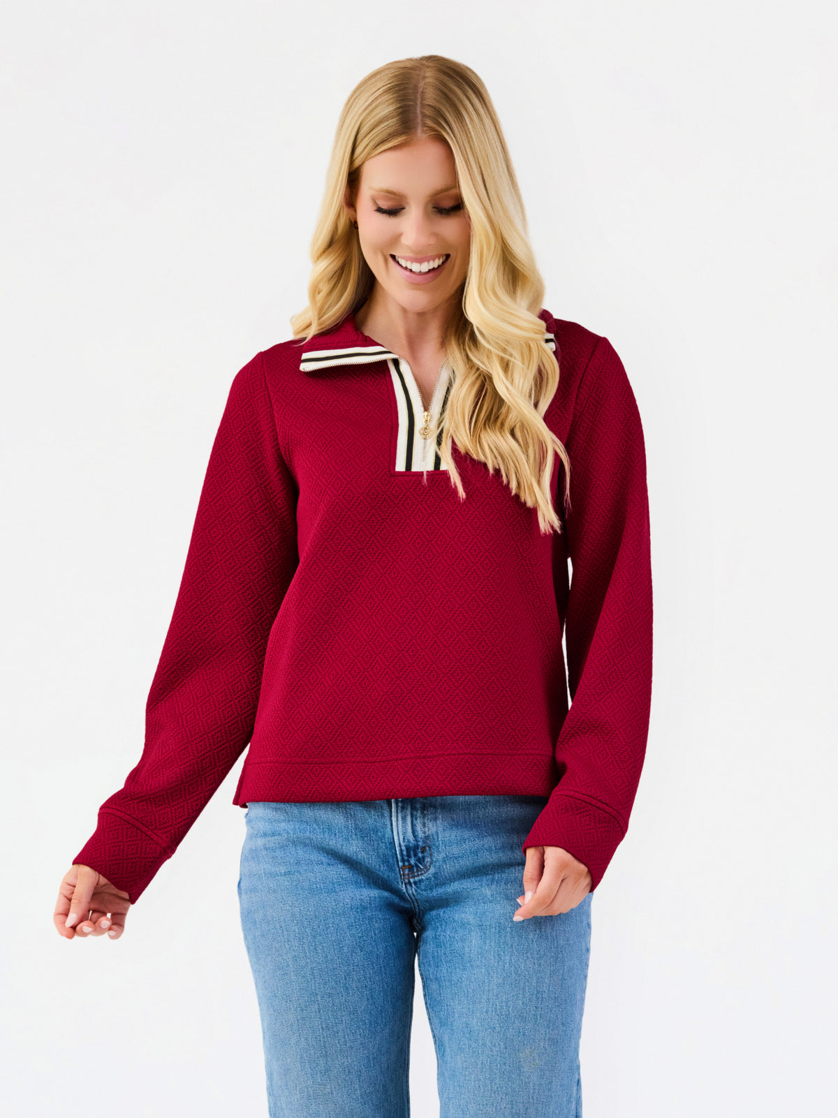 A woman wearing a burgundy long-sleeve quarter-zip sweatshirt with cream and navy striped trim on the collar, paired with blue jeans, against a plain white background.