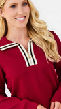 A smiling blonde woman wearing a textured deep red long-sleeve top with a white and black striped collar and a white zipper trim.