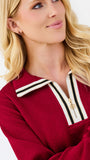 A close-up of a blonde woman wearing a textured deep red knit top with a white and black striped collar and a gold zipper.
