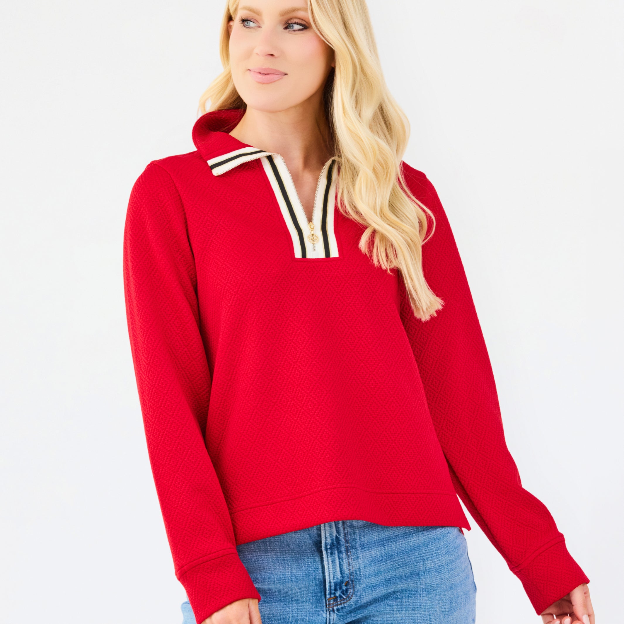 A blonde woman wearing a bright red long-sleeve knit top with a striped collar and blue jeans, standing against a plain white studio background.