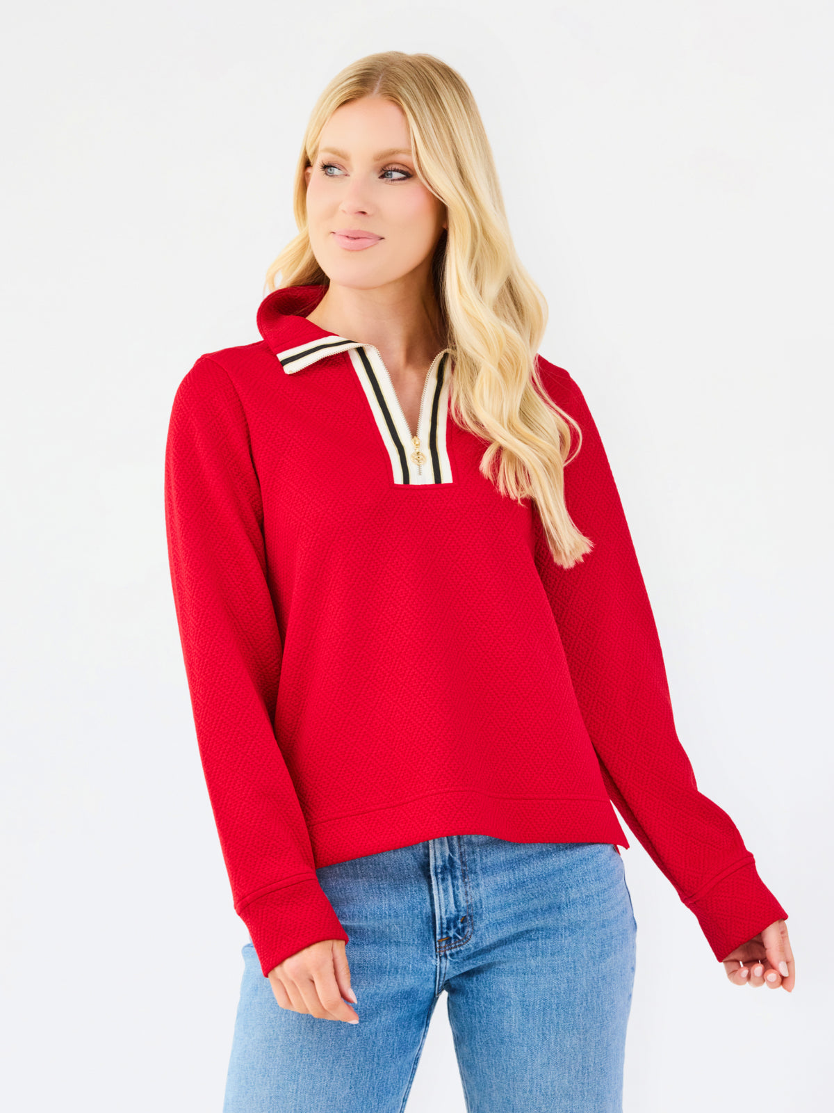 A blonde woman wearing a bright red long-sleeve knit top with a striped collar and blue jeans, standing against a plain white studio background.