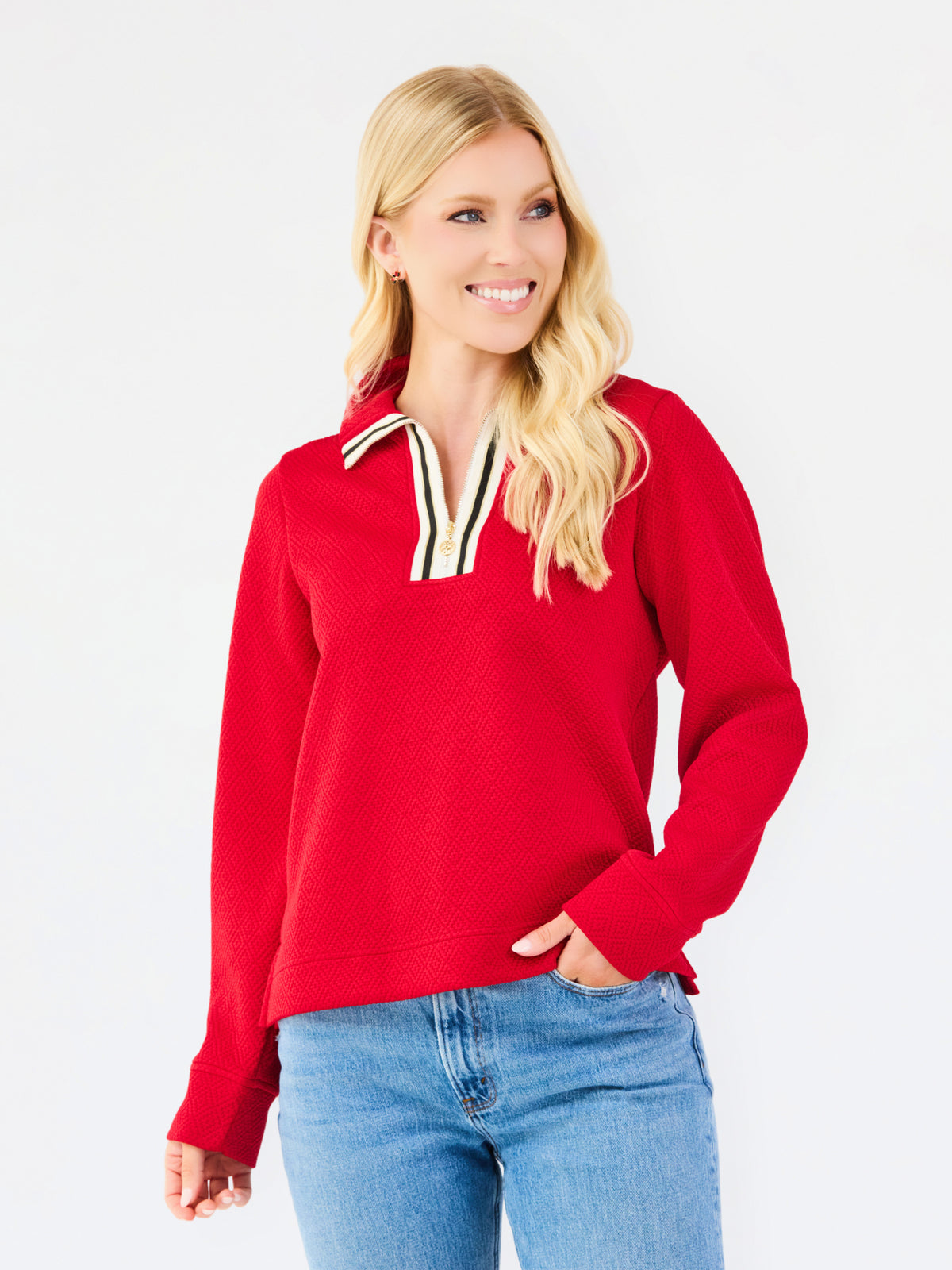 A blonde woman wearing a bright red long-sleeve knit top with a striped collar and blue jeans, standing against a plain white studio background.