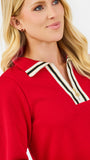 A blonde woman wearing a bright red long-sleeve knit top with a striped collar and blue jeans, standing against a plain white studio background.
