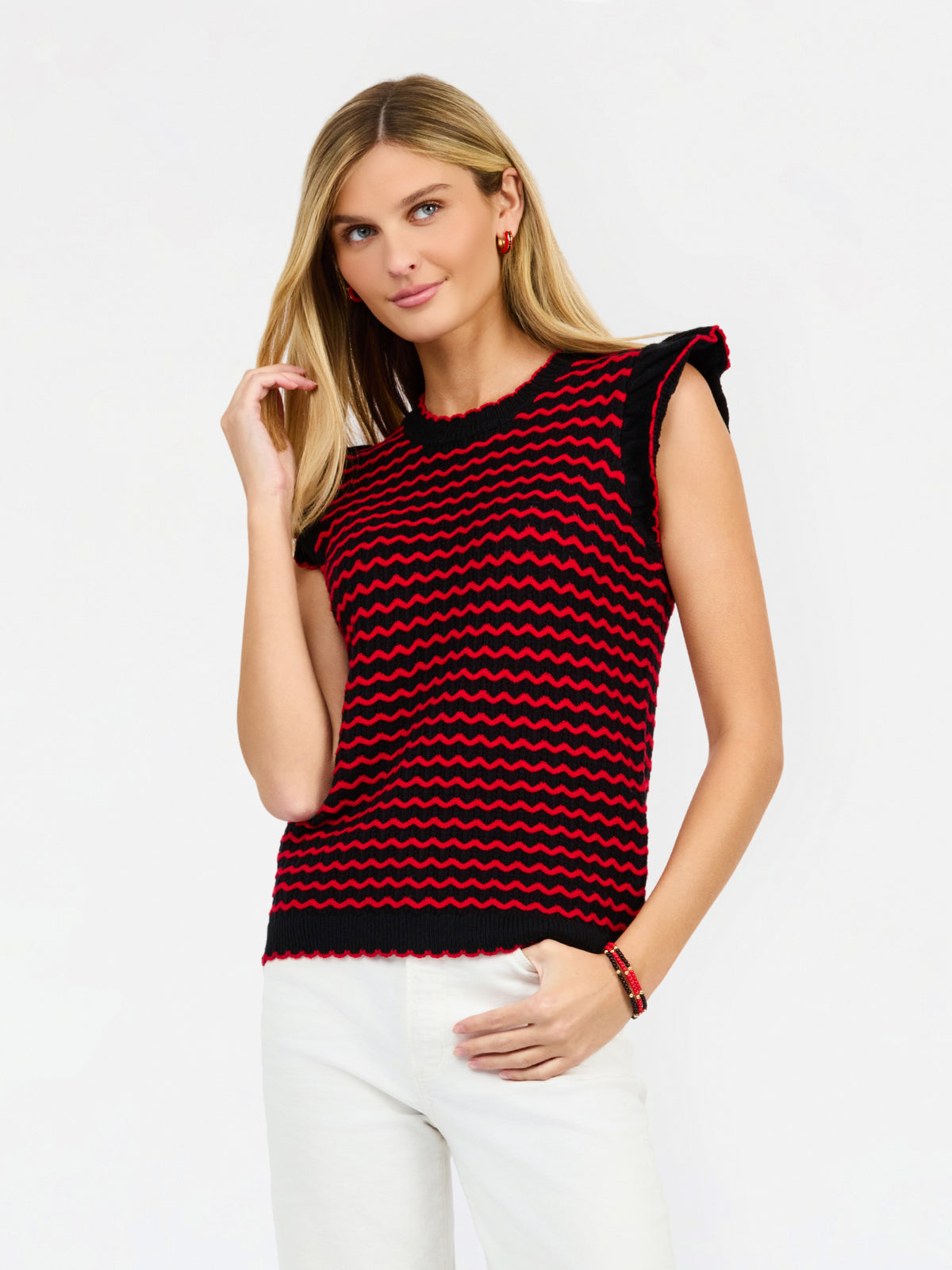 A woman modeling a sleeveless knit top with a red and black wavy stripe pattern, paired with white pants, against a plain white studio background.