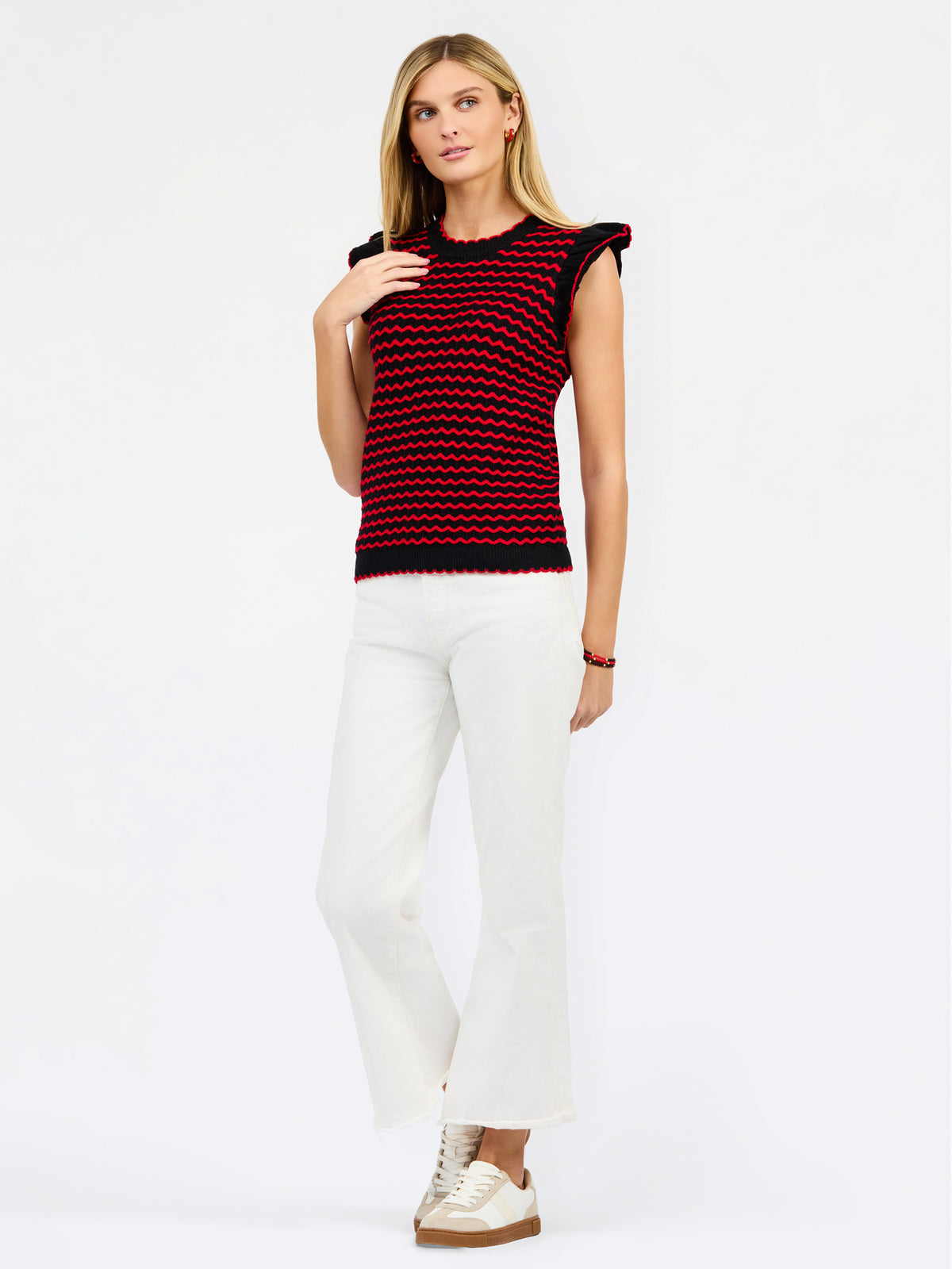 A woman modeling a sleeveless knit top with a red and black wavy stripe pattern, paired with white pants, against a plain white studio background.