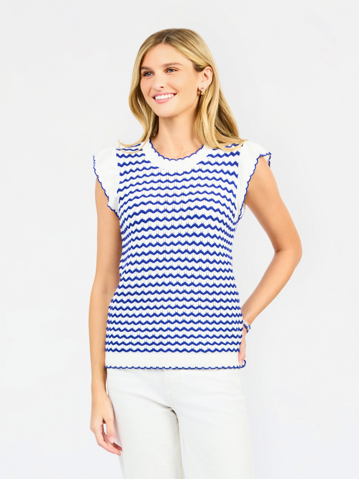 A woman modeling a sleeveless top with a blue and white wavy stripe pattern, paired with white pants, against a light gray studio background.
