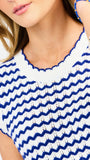 A woman modeling a sleeveless top with a blue and white wavy stripe pattern, paired with white pants, against a light gray studio background.