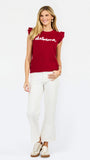 A woman modeling a red ribbed knit top with ruffled shoulder details, scalloped trim, and 'alabama' written in white script across the chest, paired with white pants against a light gray studio background.