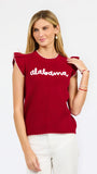 A woman modeling a red ribbed knit top with ruffled shoulder details, scalloped trim, and 'alabama' written in white script across the chest, paired with white pants against a light gray studio background.
