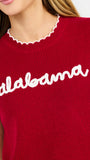 A woman modeling a red ribbed knit top with ruffled shoulder details, scalloped trim, and 'alabama' written in white script across the chest, paired with white pants against a light gray studio background.