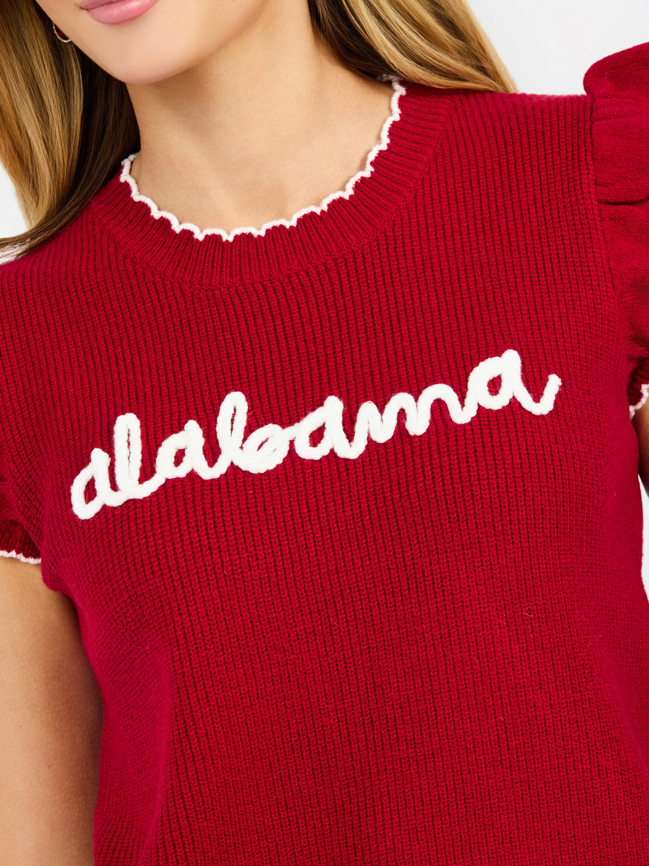 A woman modeling a red ribbed knit top with ruffled shoulder details, scalloped trim, and 'alabama' written in white script across the chest, paired with white pants against a light gray studio background.