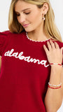 A woman modeling a red ribbed knit top with ruffled shoulder details, scalloped trim, and 'alabama' written in white script across the chest, paired with white pants against a light gray studio background.
