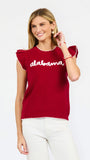 A woman modeling a red ribbed knit top with ruffled shoulder details, scalloped trim, and 'alabama' written in white script across the chest, paired with white pants against a light gray studio background.