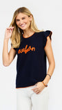 A woman modeling a navy blue ribbed knit top with orange trim and 'auburn' script across the chest, paired with white pants against a light gray studio background.