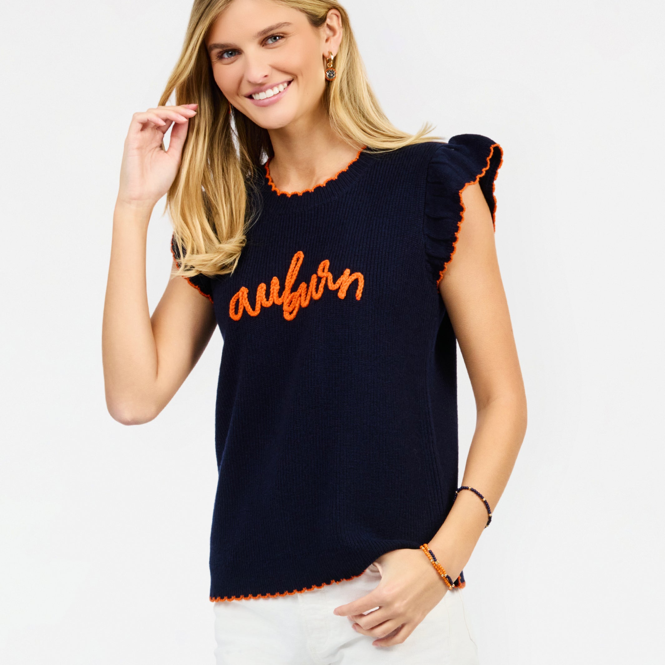 A woman modeling a navy blue ribbed knit top with orange trim and 'auburn' script across the chest, paired with white pants against a light gray studio background.